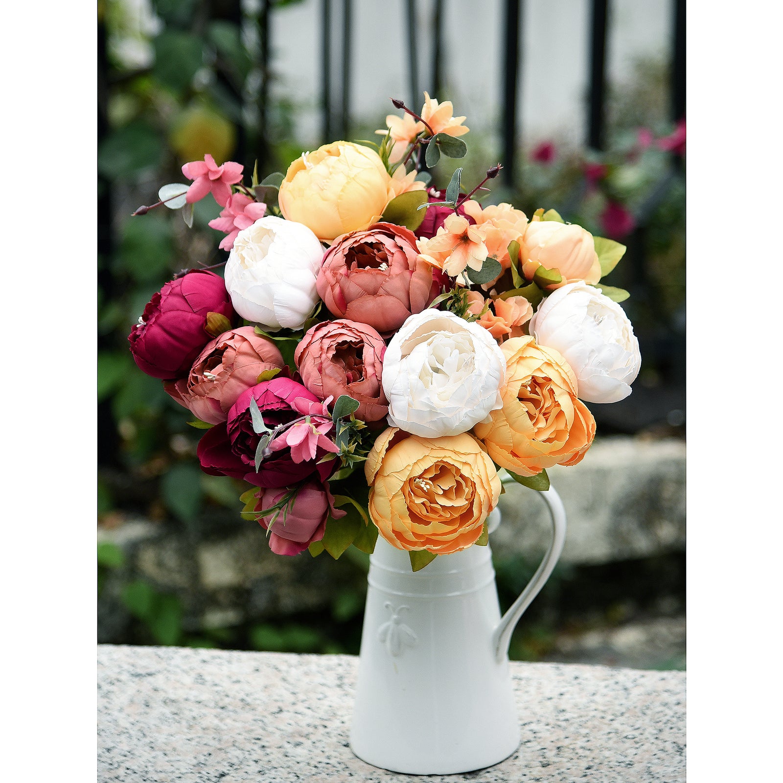 Artificial peony bouquet with cream, peach, burgundy, and pink flowers displayed in a white pitcher vase on a stone surface.