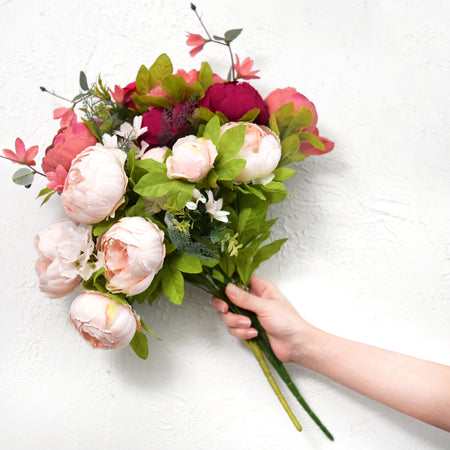 A person holding an artificial peony bouquet with mixed pink and burgundy flowers against a white background, showcasing its full and realistic appearance.