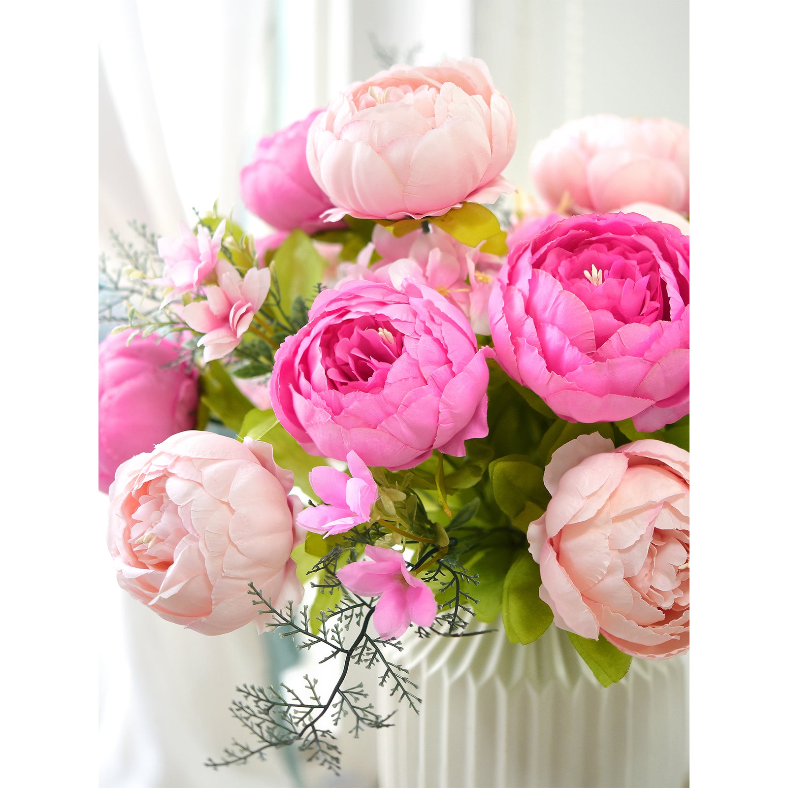 Close-up of artificial peonies in hot pink and blush shades, arranged in a white fluted vase with delicate green leaves and filler accents, beautifully styled near a sunlit window.