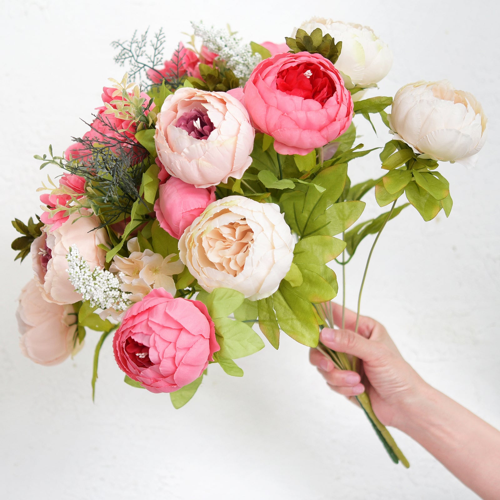 Hand holding a bunch of artificial peonies in peach, pink, and cream with greenery, showing full volume and soft texture.