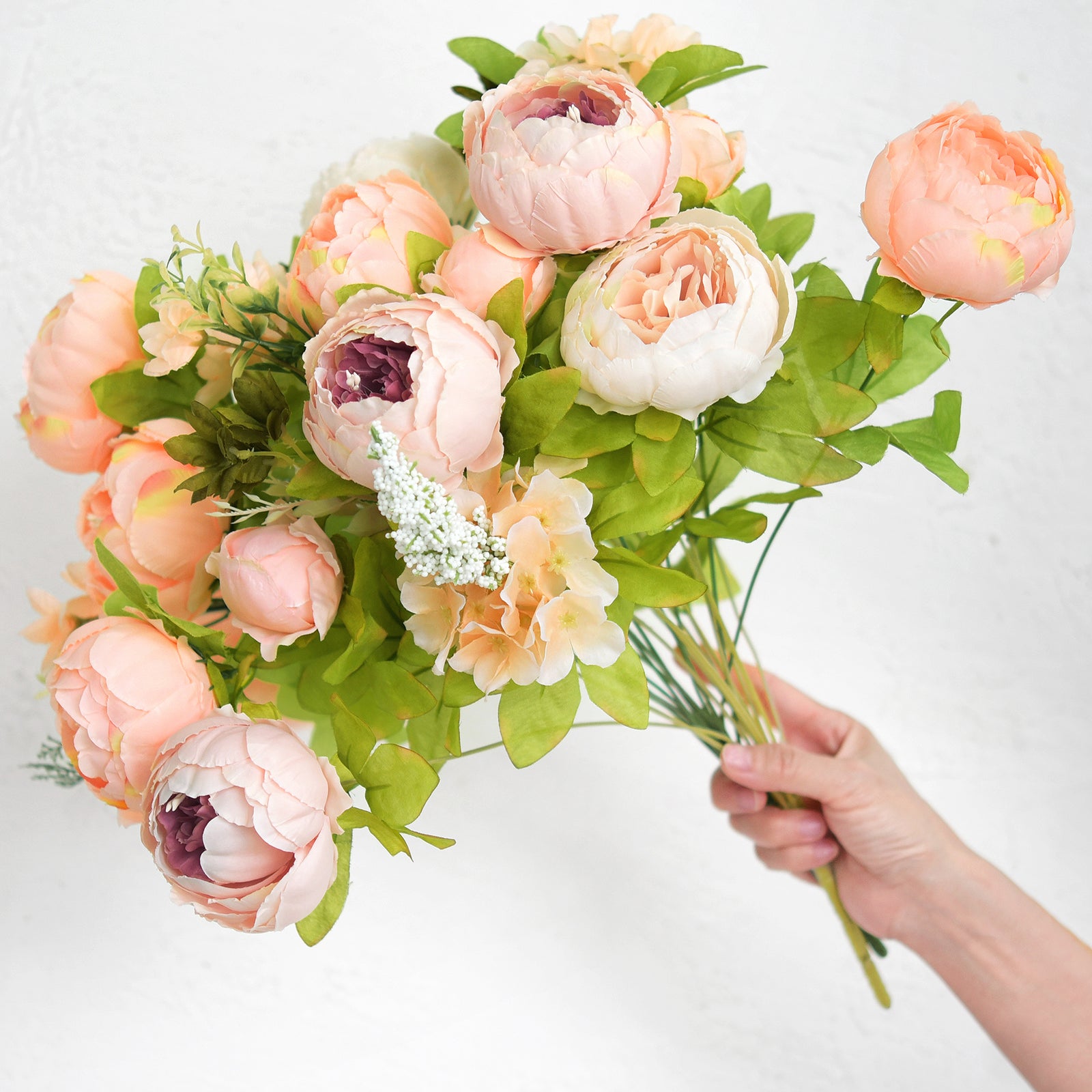 Hand holding a bunch of artificial peonies with full volume and mix of blush and peach hues, ready for DIY floral styling.