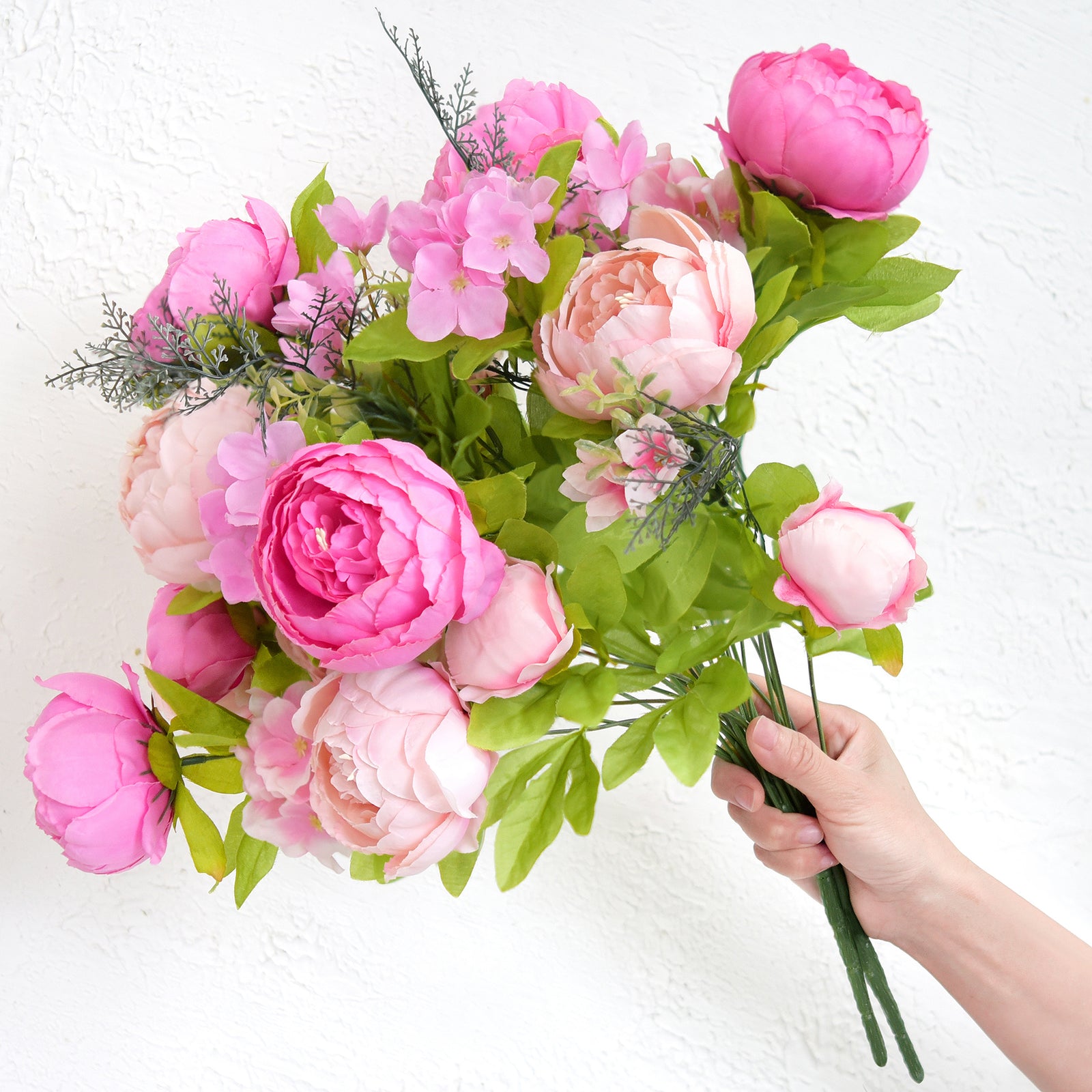 Person holding a bunch of hot pink and blush artificial peonies with greenery, showing lifelike detail and bouquet fullness.