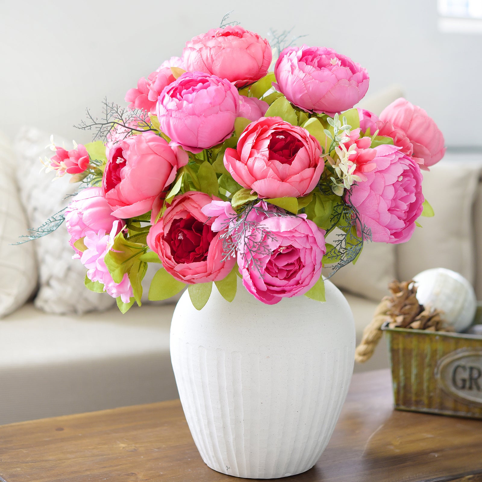 Lifelike artificial peonies in bold pinks arranged in a white vase, placed on a wooden coffee table with a candle and pinecones for cozy interior styling.