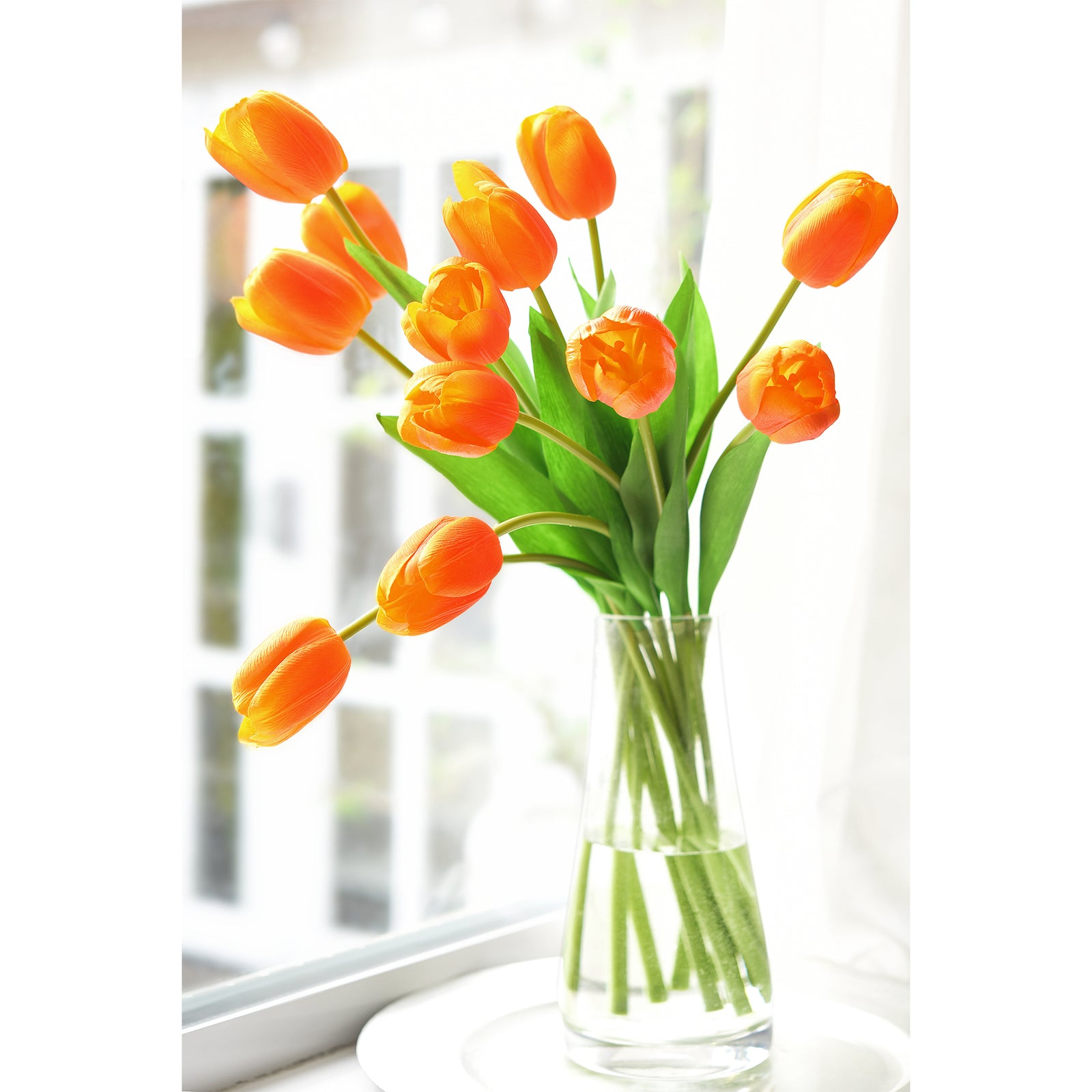 Orange artificial tulips placed in a glass vase near a sunny window for spring décor.