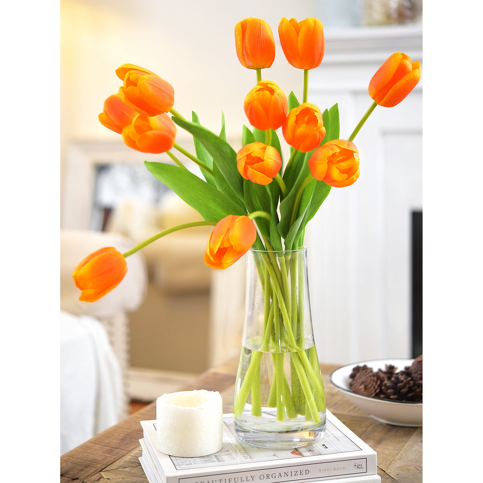 Artificial orange tulips arranged in a glass vase on a cozy living room table with books and candles.