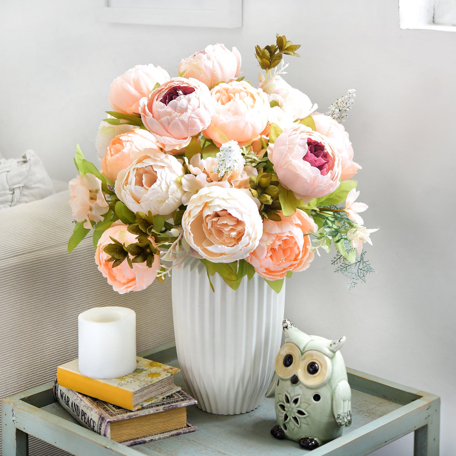 Artificial peach and cream peonies in a white vase with owl figurine and candle on a glass side table, styled for cozy living spaces or seasonal decor.