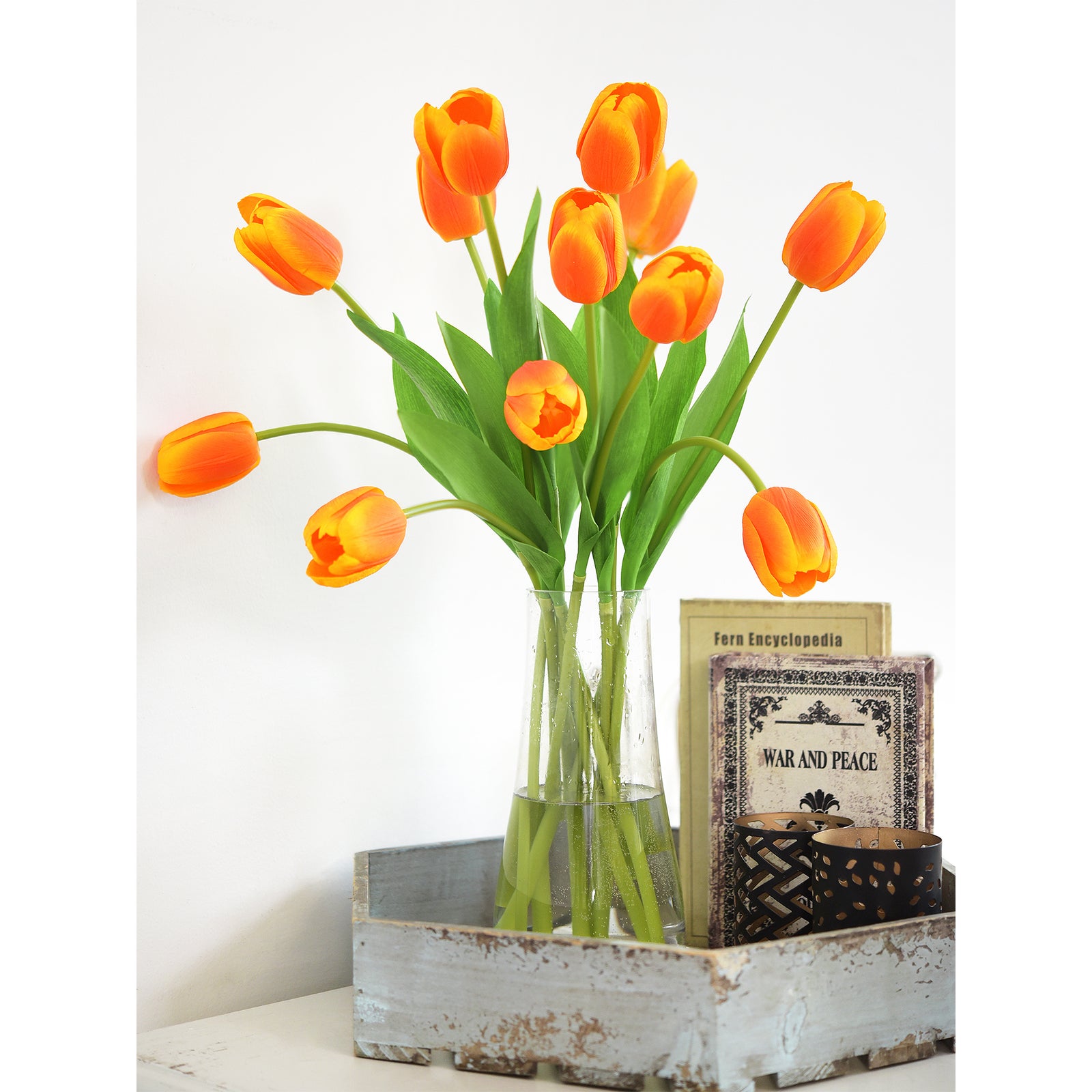 Long-stem orange tulips in a glass vase on a rustic tray with candles and books.