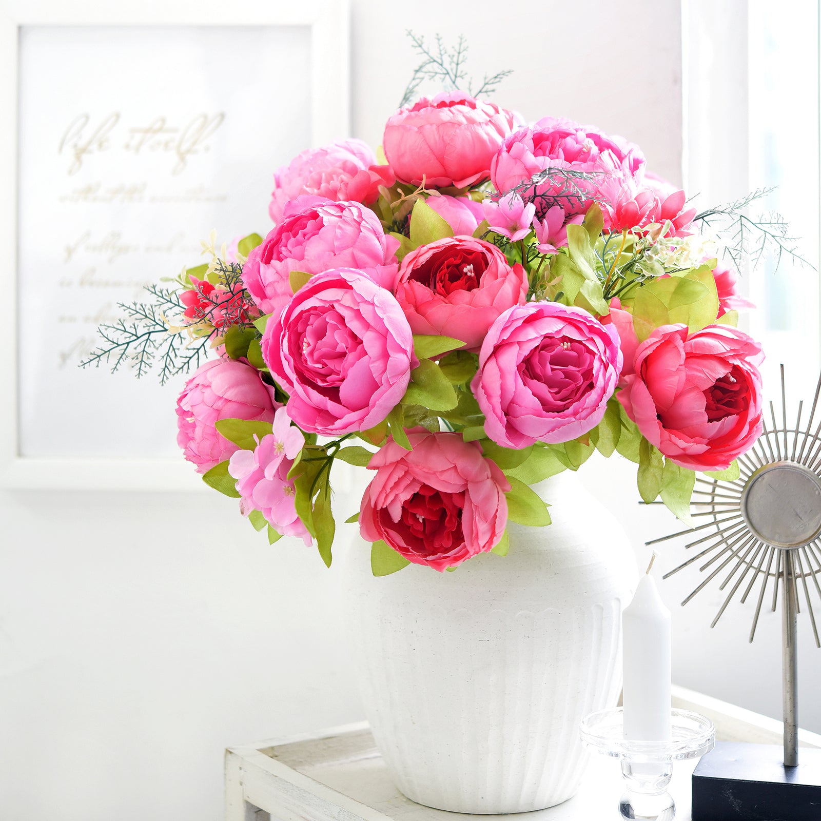 Vibrant pink artificial peony bouquet in a fluted vase placed on a white cabinet near framed wall art and sunlight, ideal for bright interior spaces.
