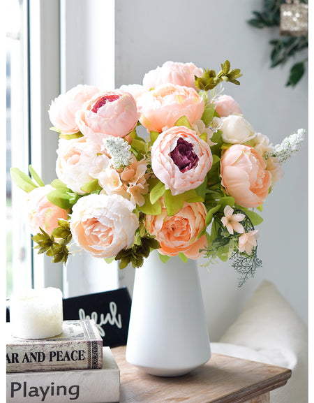 Elegant silk peony bouquet in a modern white vase displayed on a table by a window, featuring light peach and cream blooms with realistic greenery.