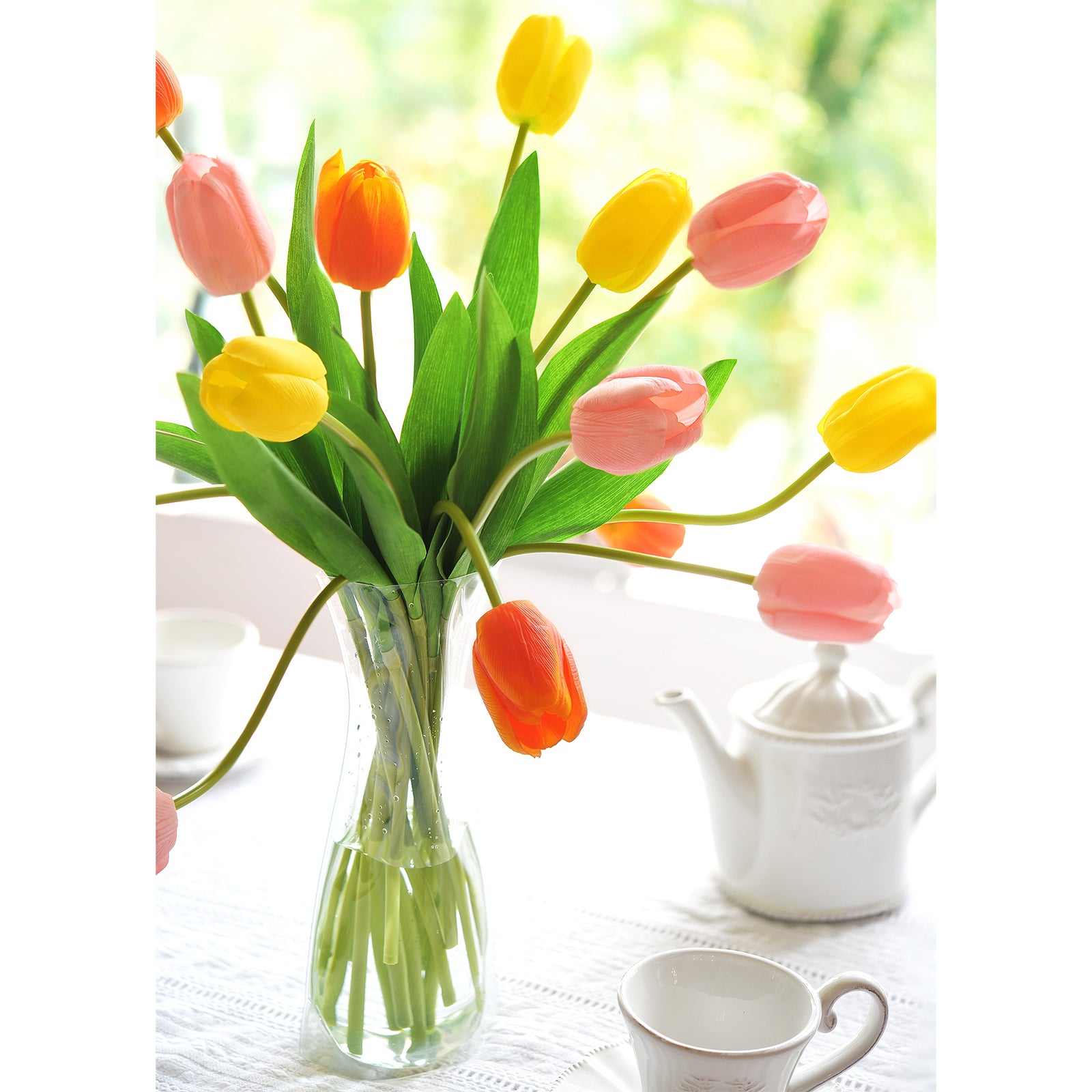 Vibrant artificial tulip bouquet in orange, yellow, and pink displayed in a transparent vase on a table, surrounded by white teaware and natural sunlight in the background.