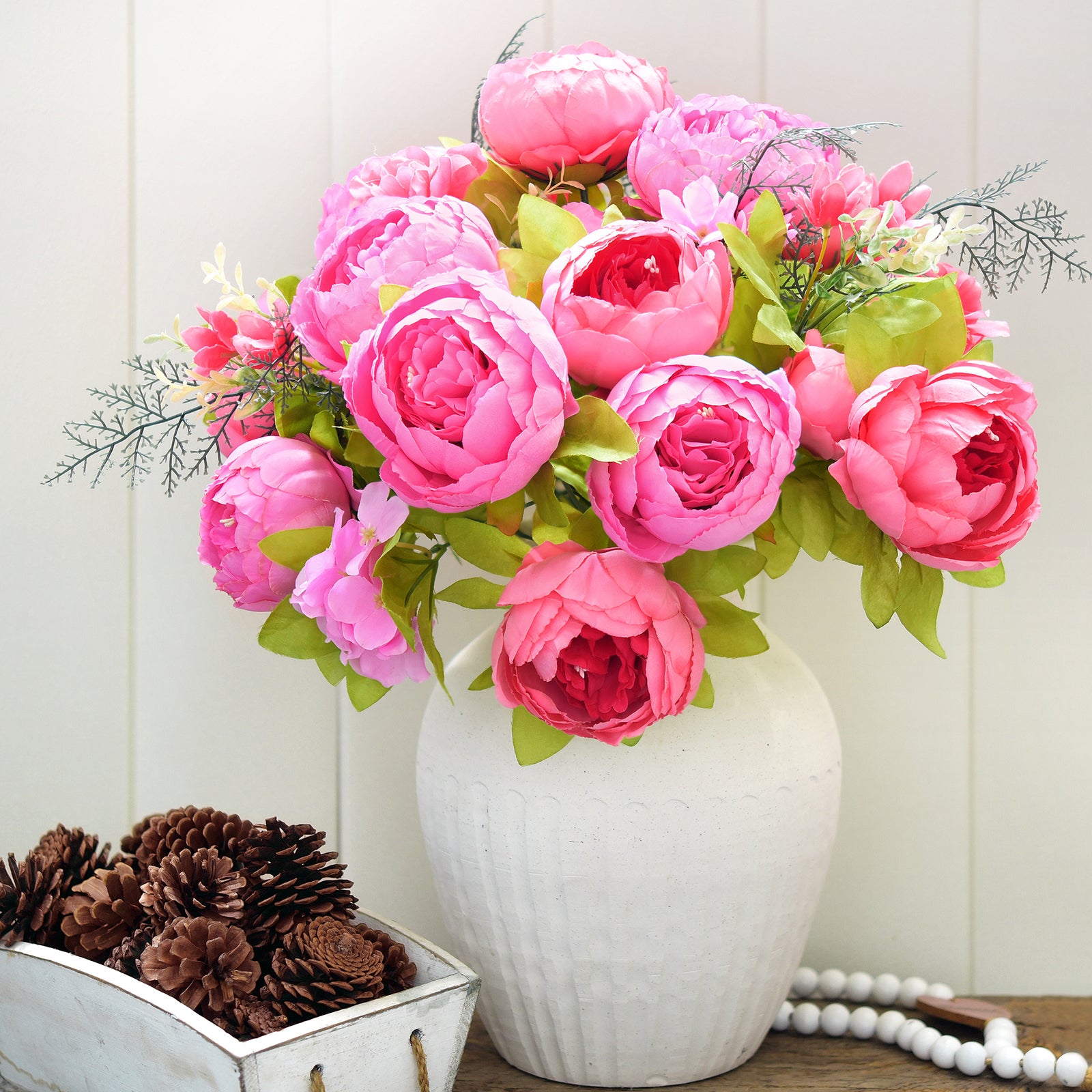 Hot pink artificial peony bouquet displayed in a white ceramic vase alongside pearl accents and pinecones, styled for warm seasonal décor.