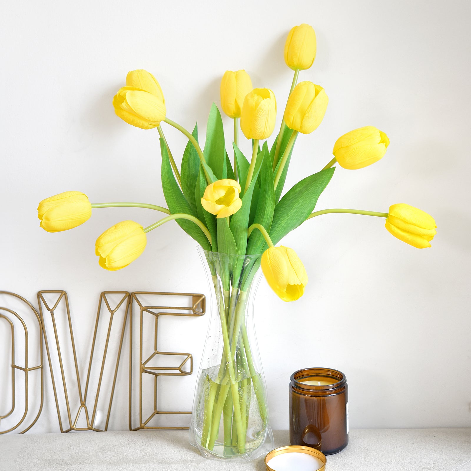 Artificial yellow tulip bouquet arranged in a vase with cozy home decor, including a gold "LOVE" sign and a candle.