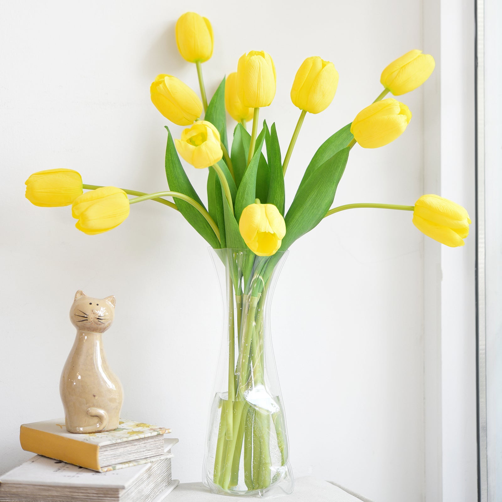 Vibrant yellow artificial tulips displayed in a tall glass vase on a minimalist side table with books and a ceramic cat figurine.