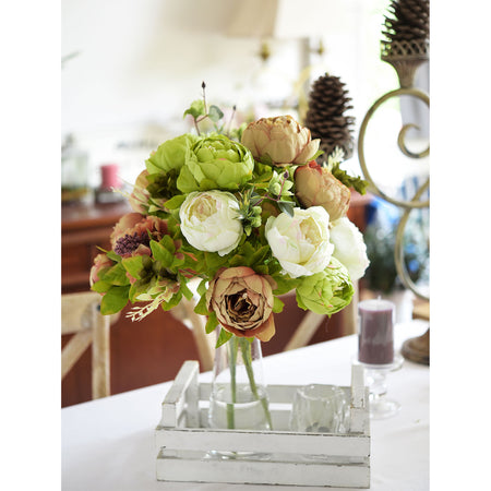 An artificial green and white peony bouquet arranged in a clear glass vase on a table, adding a fresh, elegant touch to the room decor.